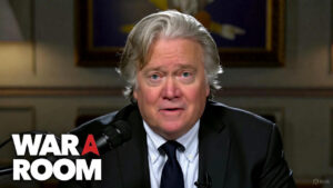 Steve Bannon speaking at War Room broadcast desk with American flag and political graphics in background